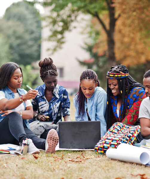 Group of five african college students spending time together on campus at university yard. Black afro friends sitting on grass and studying with laptops.